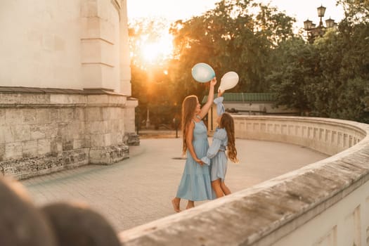 Daughter mother run holding hands. In blue dresses with flowing long hair, they hold balloons in their hands against the backdrop of a sunset and a white building