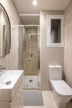 Modern compact beige tiled bathroom with glass walk-in shower, sink and mirror and white toilet with window. Concept of a tiny bathroom in a hotel or small apartment.