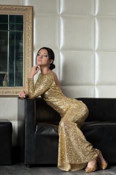 Full length portrait of young brunette woman in gold dress posing on sofa