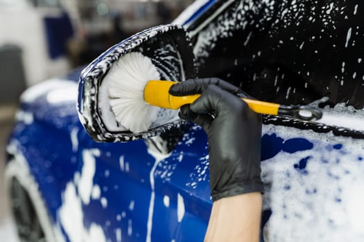 Hand brush washing of car side mirror with foam in car detailing service. Car wash worker washes auto body