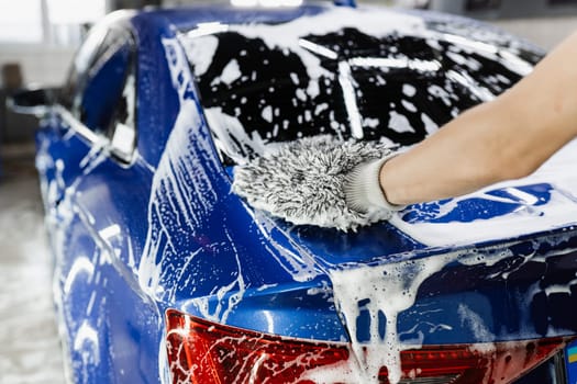 Car washer doing manual foam washing in auto detailing service. Hand washing with microfiber glove with foam car body in garage