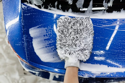 Car washer doing manual foam washing in auto detailing service. Hand washing with microfiber glove with foam car body in garage