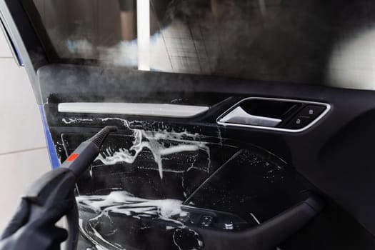 Steam vaping cleaning inside handle of car door after foam washing with brush. Worker in car detailing service washing leather interior of clients auto using steam generator