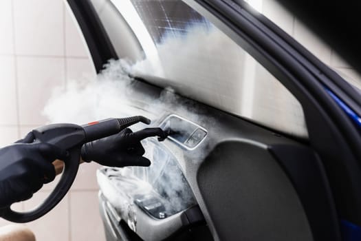 Steam vaping cleaning inside handle of car door after foam washing with brush. Worker in car detailing service washing leather interior of clients auto using steam generator