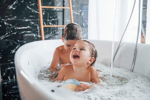Two kids having fun and washing themselves in the bath at home. Helping each other.
