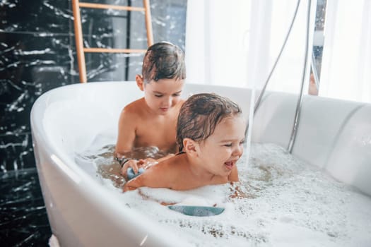Two kids having fun and washing themselves in the bath at home. Helping each other.
