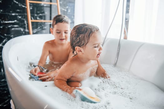 Two kids having fun and washing themselves in the bath at home. Helping each other.