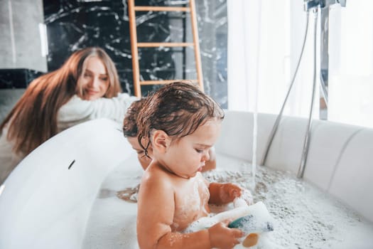 Young mother helps her son and daughter. Two kids washing in the bath.