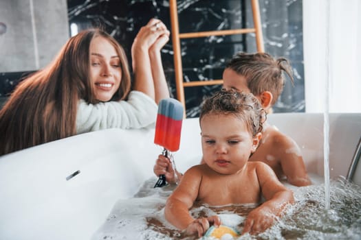 Young mother helps her son and daughter. Two kids washing in the bath.