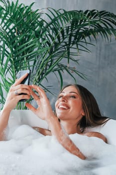 Uses phone. Beautiful young woman have free time at home in the bath.