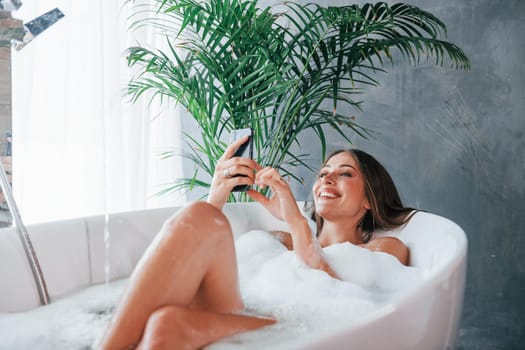 Uses phone. Beautiful young woman have free time at home in the bath.