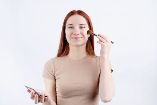 Joyful redhead young woman using makeup brush and blusher isolated on white background, copy space, natural beauty