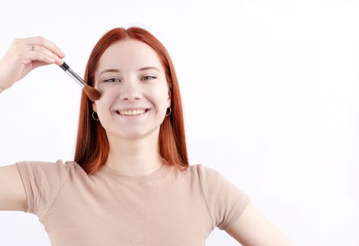 Joyful redhead young woman using makeup brush making up isolated on white background, copy space, natural beauty