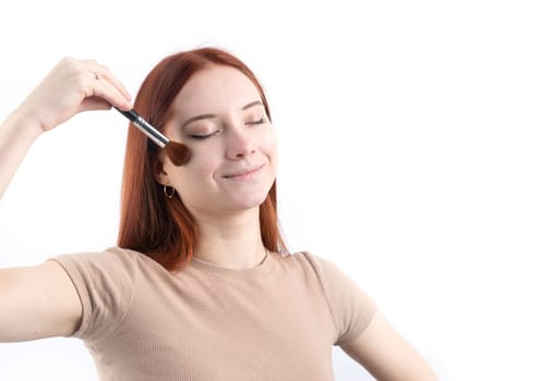 Joyful redhead young woman using makeup brush making up isolated on white background, copy space, natural beauty