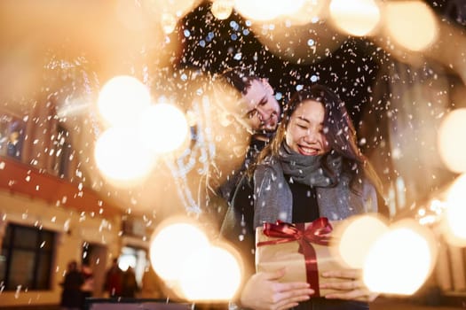 Holds gift box. Happy multiracial couple together outdoors in the city celebrating New year.