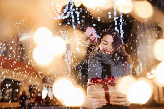 Holds gift box. Happy multiracial couple together outdoors in the city celebrating New year.