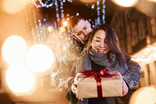 Holds gift box. Happy multiracial couple together outdoors in the city celebrating New year.