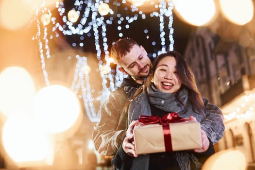 Holds gift box. Happy multiracial couple together outdoors in the city celebrating New year.
