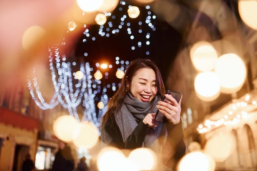 Uses phone. Cute and happy asian young woman outdoors in the city celebrates New year.