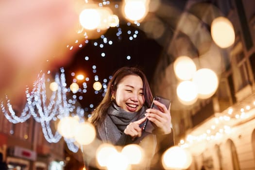Uses phone. Cute and happy asian young woman outdoors in the city celebrates New year.