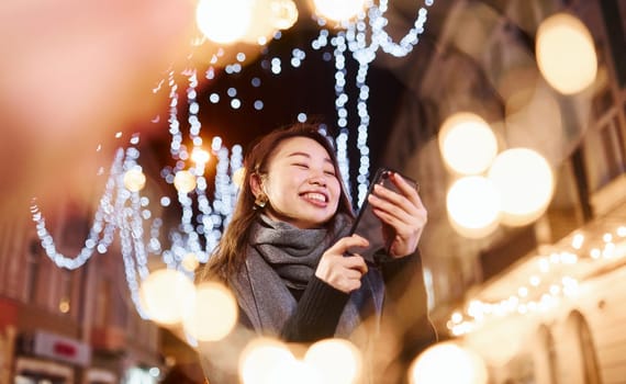 Uses phone. Cute and happy asian young woman outdoors in the city celebrates New year.