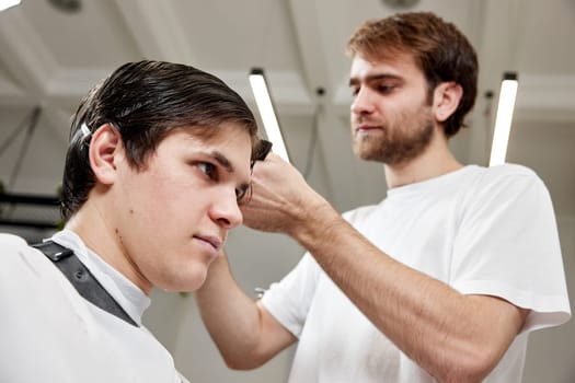 professional male hairstylist serving young caucasian client man at modern barber shop.