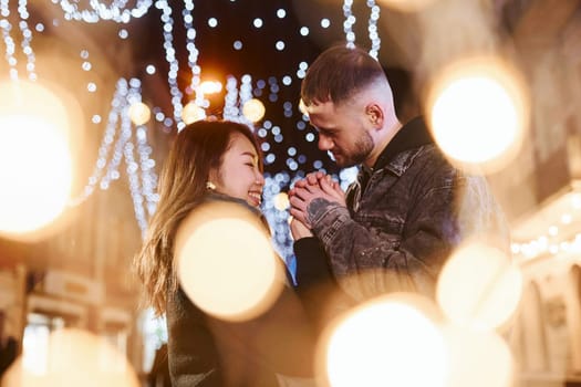 Artificial lighting by garlands. Happy multiracial couple together outdoors in the city celebrating New year.