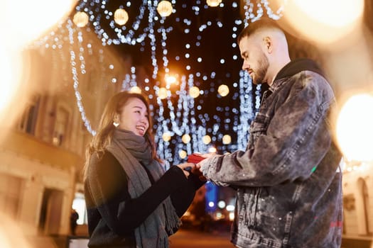 Holds gift box. Happy multiracial couple together outdoors in the city celebrating New year.
