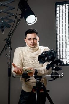 surprised caucasian photographer with digital camera on tripod holding cash on gray background with lighting equipment