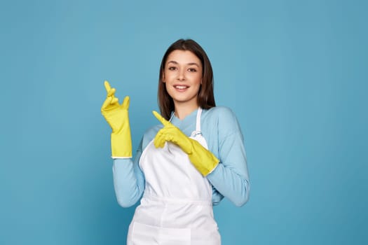brunette caucasian woman in yellow rubber gloves pointing fingers aside at empty space isolated on blue background.