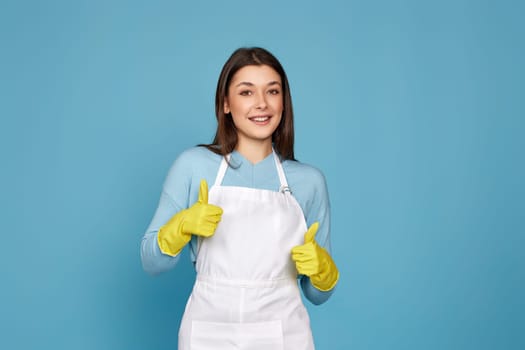 beautiful brunette woman in yellow rubber gloves and cleaner apron showing ok sign on blue background.