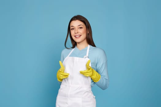 brunette woman in yellow rubber gloves and cleaner apron pointing fingers to camera isolated on blue background.