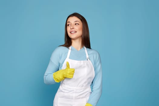 beautiful brunette woman in yellow rubber gloves and cleaner apron showing ok sign on blue background.