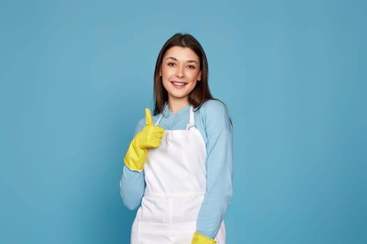 beautiful brunette woman in yellow rubber gloves and cleaner apron showing ok sign on blue background.