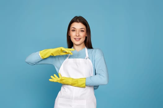 beautiful brunette woman in yellow rubber gloves and cleaner apron showing measure symbol gesture on blue background. cleaning