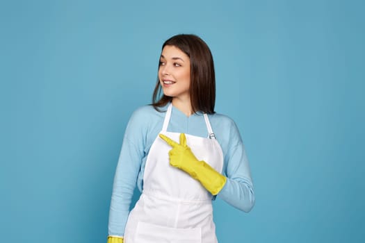 brunette caucasian woman in yellow rubber gloves pointing fingers aside at copyspace isolated on blue background.