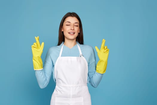 beautiful brunette caucasian girl in yellow rubber gloves with finger crossed gesture isolated on blue background.