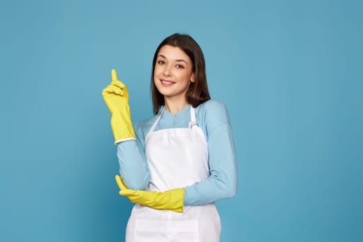 pretty smiling woman in rubber gloves and apron holding index finger up with new idea and looking at camera on blue background. cleaning