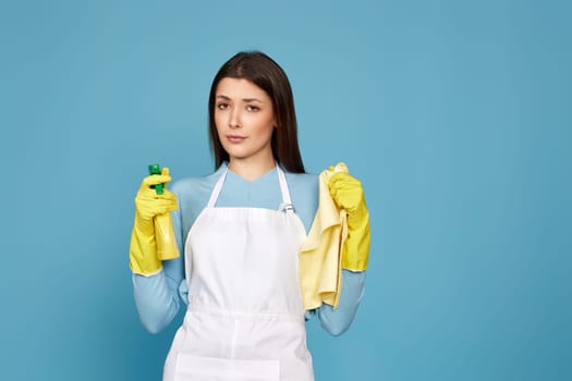 skeptic and nervous, frowning woman in gloves and cleaner apron with cleaning rag and detergent sprayer on blue background.