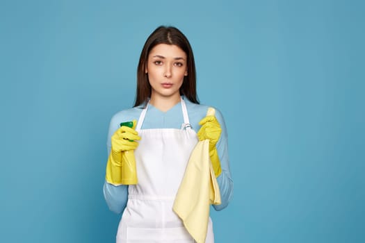 caucasian woman in rubber gloves and cleaner apron with cleaning rag and detergent sprayer on blue background.