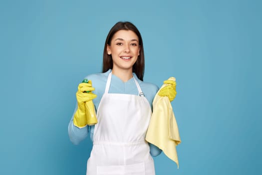 smiling caucasian woman in yellow rubber gloves and cleaner apron with cleaning rag and detergent sprayer on blue background.
