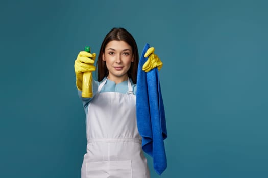 pretty girl in rubber gloves and cleaner apron holding cleaning rag and detergent sprayer on blue background. focus on hands