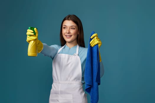 happy young woman in rubber gloves and cleaner apron holding cleaning rag and detergent sprayer on blue background. focus on hands