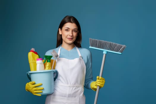 beautiful girl in yellow rubber gloves and cleaner apron holding bucket of detergents and broom on blue background.