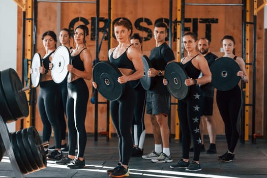 Holding weights. Group of young sportive people have crossfit day indoors in gym.