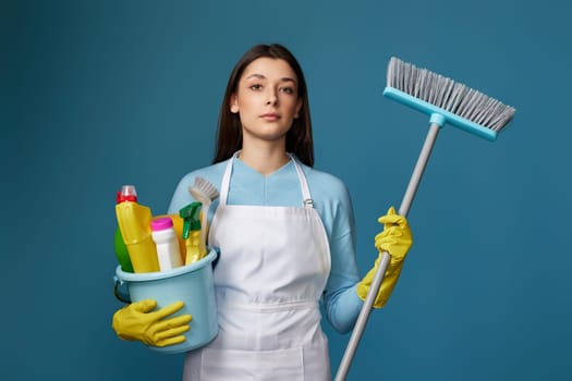 pretty calm woman in yellow rubber gloves and cleaner apron holding bucket of detergents and broom on blue background.