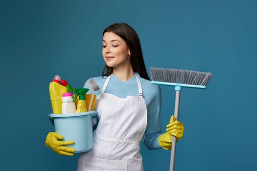 smiling woman in yellow rubber gloves and cleaner apron holding bucket of detergents and broom on blue background.