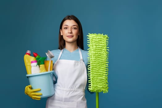Attractive woman in yellow rubber gloves and cleaner apro n holding bucket of detergents and mop on blue background.