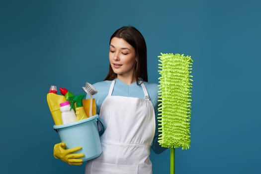 pretty young woman in yellow rubber gloves and cleaner apron holding bucket of detergents and mop on blue background.