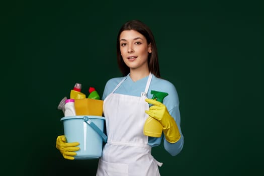 beautiful woman in yellow rubber gloves and cleaner apron holding bucket with cleaning supplies and detergent sprayer on dark green background.